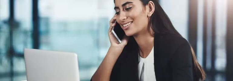 Woman talking on a mobile phone while using a laptop in an office setting.