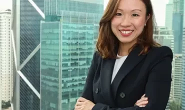 Smiling woman in business attire standing by office window with city skyscrapers in the background.