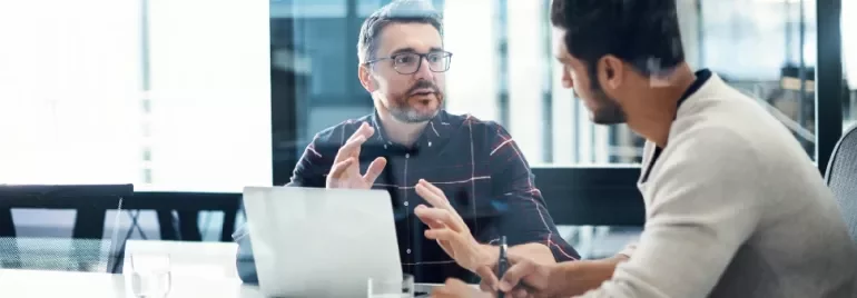 Two men having a discussion in an office with a laptop open on the table.