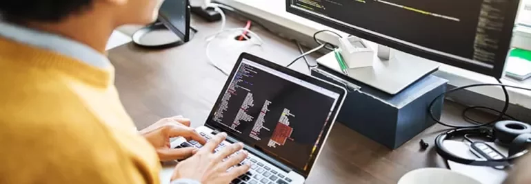 Person coding on a laptop with a large monitor in the background.