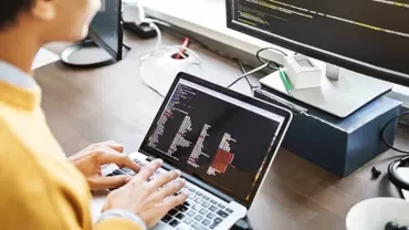 Person coding on a laptop with a large monitor in the background.