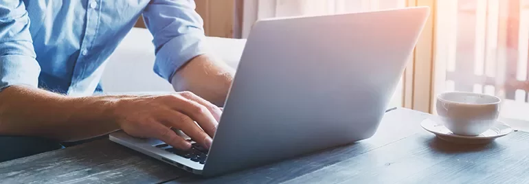 Person typing on a laptop at a wooden desk with a cup of coffee.