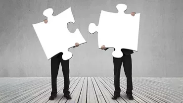Two people holding large blank puzzle pieces standing on a wooden floor.
