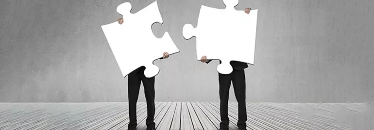 Two people holding large blank puzzle pieces standing on a wooden floor.