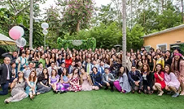 Large group of people posing outdoors on a lawn with trees and flowers.