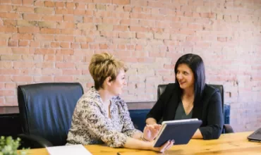 Two women having a discussion at a wooden table in an office setting.