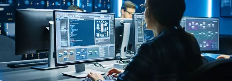 A person working at a computer with multiple screens displaying complex data and charts in a tech office.
