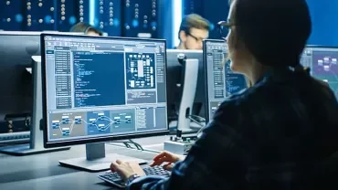 A person working at a computer with multiple screens displaying complex data and charts in a tech office.