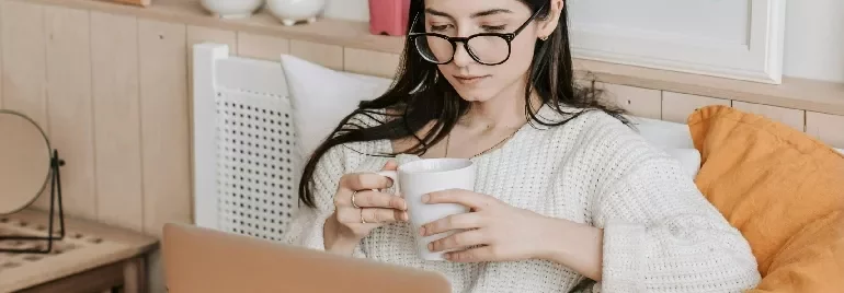 Woman with glasses holding a mug and using a laptop in a cozy room.