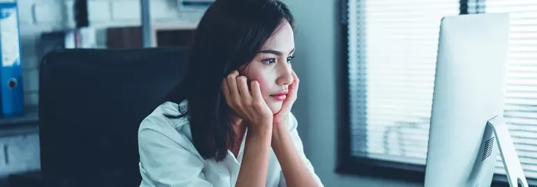 Woman sitting at a desk looking at her computer screen thoughtfully.