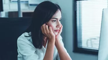 Woman sitting at a desk looking at her computer screen thoughtfully.