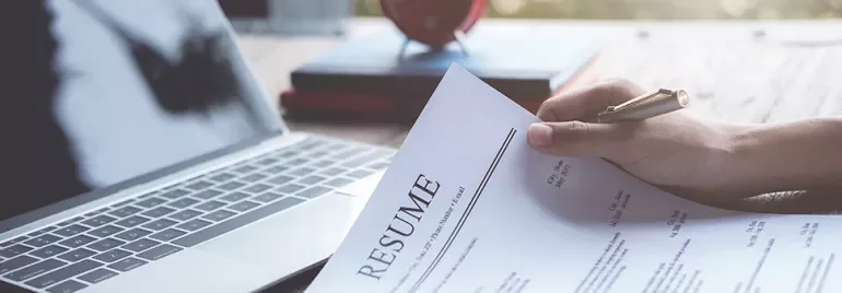 Person holding a resume in front of a laptop on a wooden desk.