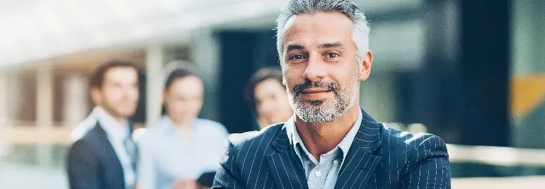Smiling man in a suit standing confidently with blurred people in the background.