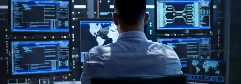 Man in front of multiple screens showing data in a dark server room.