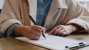 Person in a beige coat writing on a document at a wooden table.