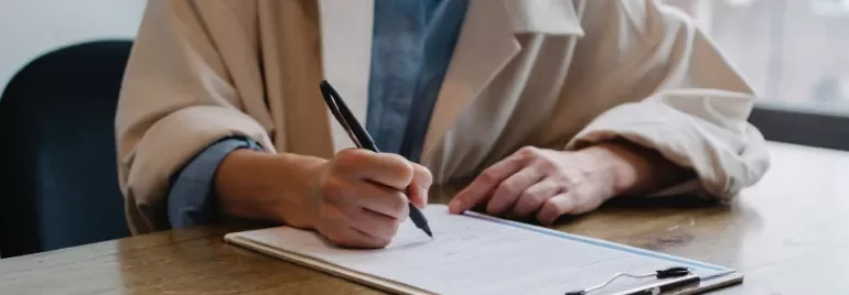 Person in a beige coat writing on a document at a wooden table.