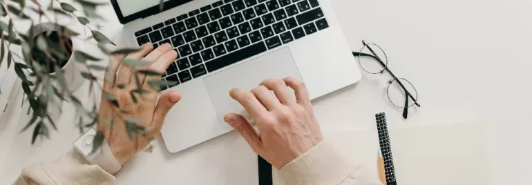 Person typing on a laptop with notebook and glasses on a desk.