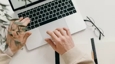 Person typing on a laptop with notebook and glasses on a desk.